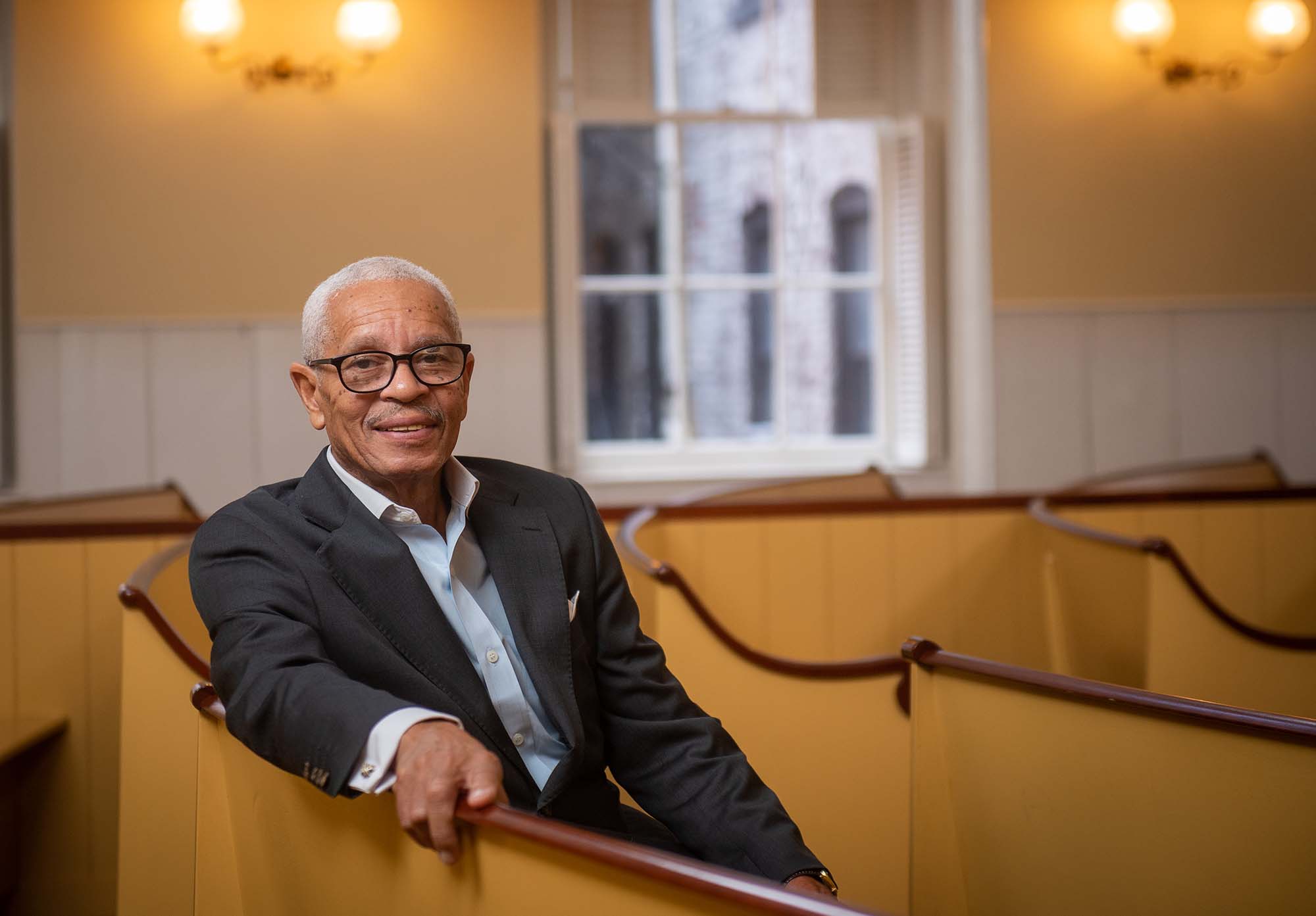 Photo of MET alum Leon Wilson, President and Chief Executive Officer at the Museum of African American History (MAAH) sitting in a pew at the meeting house on November 18, 2021. Wilson is an older Black man with short, graying hair and large black-rimmed glasses, who smiles, wears a suit and rests his right arm on the wooden, painted pews.
