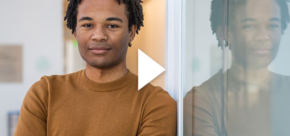 Photo of Ethan Strauther, a young Black man with short dreads wearing a burnt yellow sweater. He leans against a glass wall that reflects his image back. White play button is overlaid on top