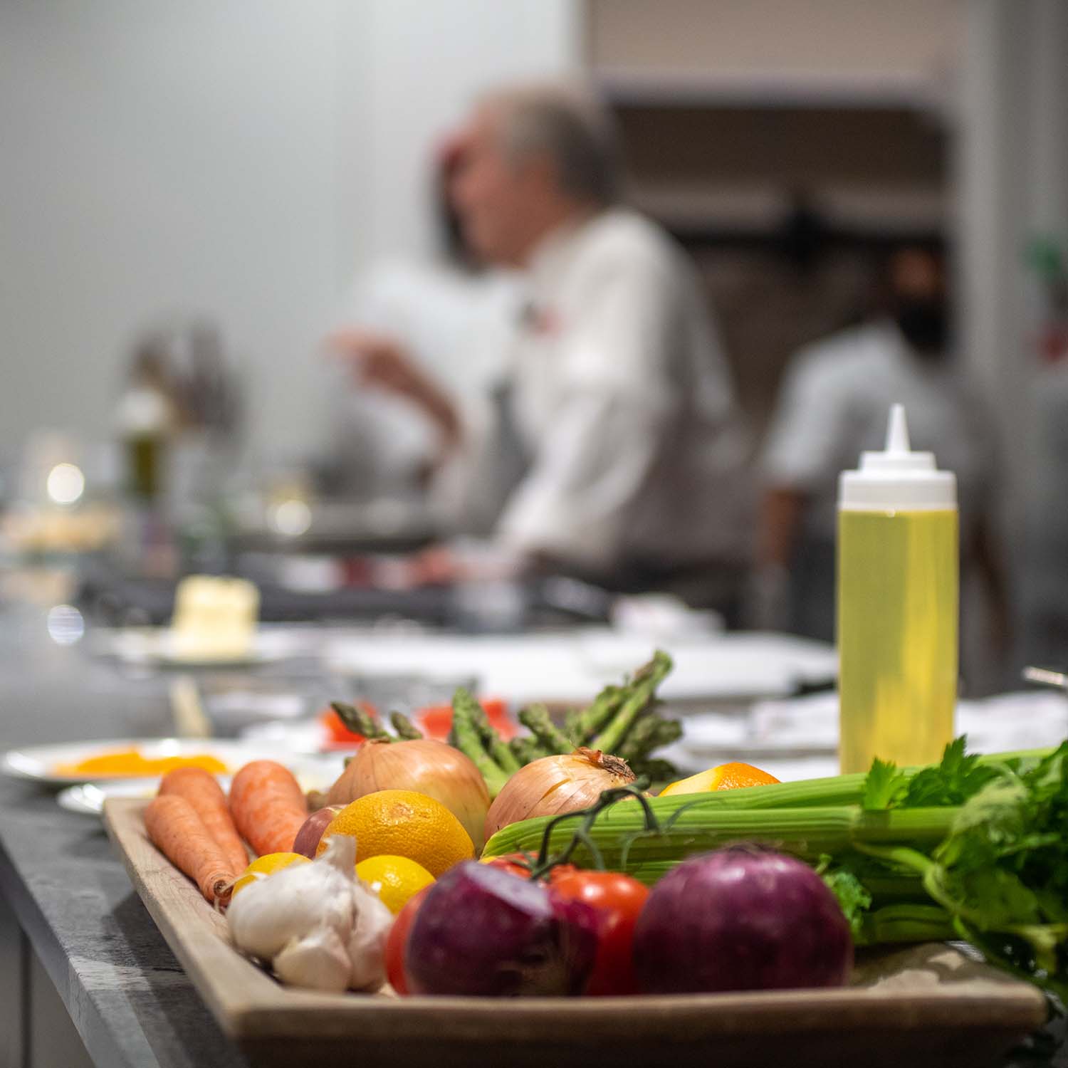 Photo of a wooden tray filled with vegetables Pepin will chop on the demo kitchen counter, with Pepin seen blurred in the background. Vegetable son the tray include garlic, celery, onions, carrots and lemons.