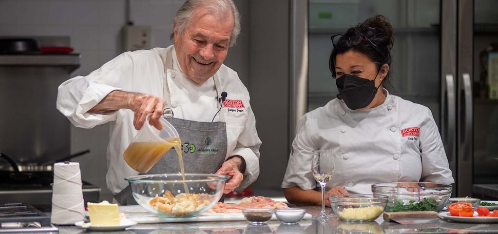 Photo of Jacques Pépin (Hon.’11), assisted by Lisa Falso-Doherty, as he pours stock into the bread and spinach stuffing for chicken ballottine in the newly renovated Groce-Pépin Culinary Innovation Lab kitchen. Pépin wears a gray Pépin foundation apron and chef jacket with BU logo, and Falso-Doherty also wears a chef jacket with the BU logo and wears a black mask. Bowls of ingredients rest on the counter before them.