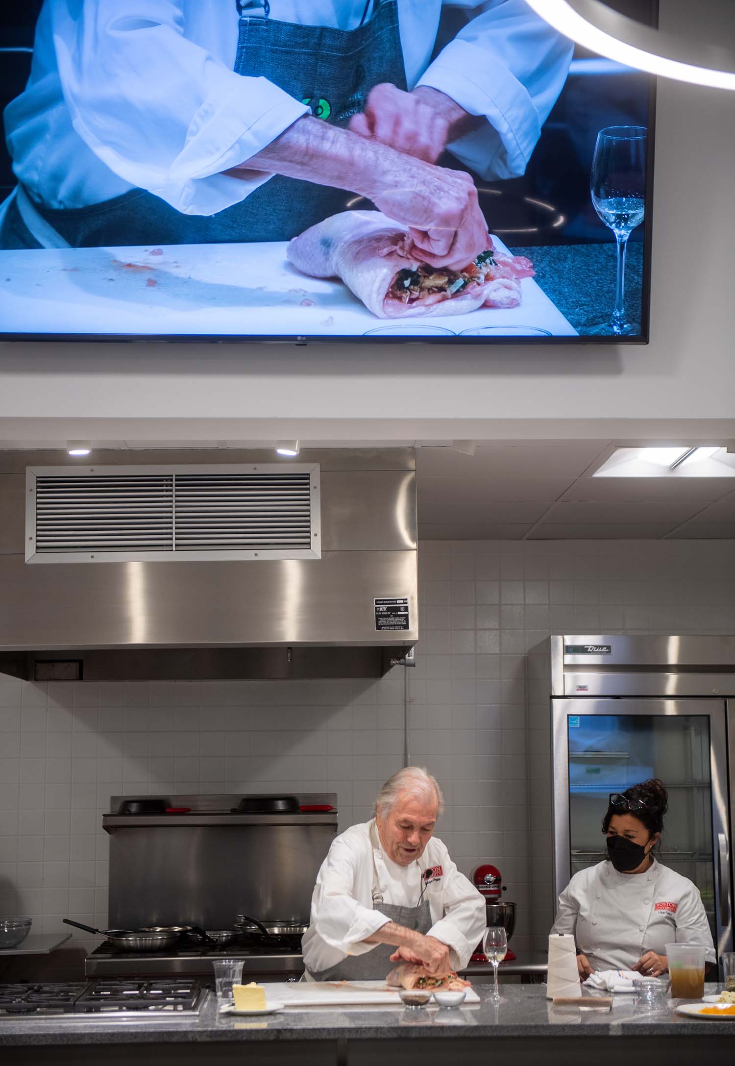 Photo of Jacques Pépin, with Lisa Falso-Doherty to his left, as he wraps and ties the stuffed chicken ballottine in the new demo kitchen. Above him, a monitor zooms in on his hands as he folds over the chicken.