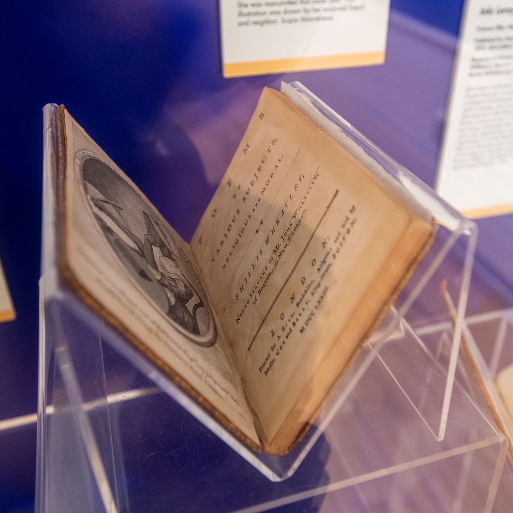Photo of an old book poems on Various Subjects, Religious and Moral by Phillis Wheatley on display at Museum of African American History (MAAH) on Joy Street in Boston. The pages are yellowed with age. An engraving of a young African American woman writing is seen on the left page.