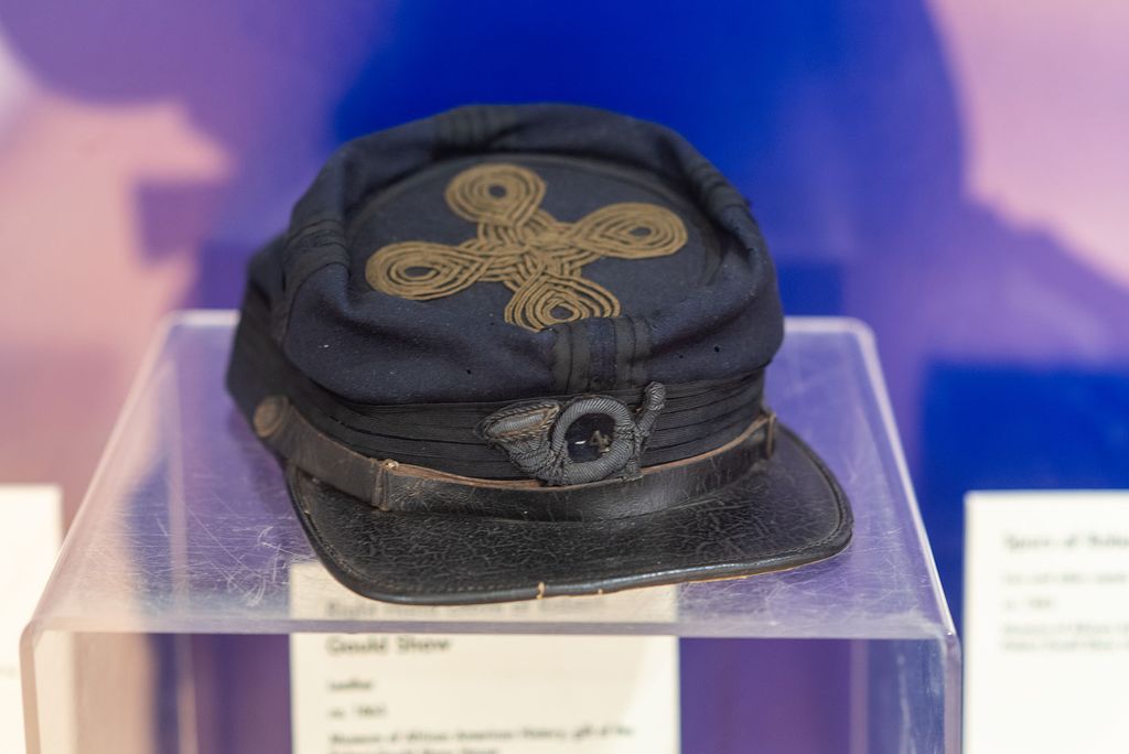Photo of the hat of Colonel Robert Gould Shaw on display at Museum of African American History (MAAH) on Joy Street in Boston. The hat is blue with a black rim, with a gold cross shape embroidered on the top.