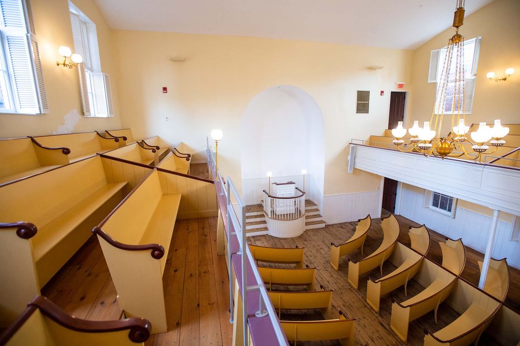 Photo of the inside of the Meeting House at the Museum of African American History (MAAH) on Joy Street in Boston. The walls are painted a light yellow, and the wooden pews are painted a mustard yellow color. The photo is taken on the second floor balcony, and you can see the white pulpit at the front of the house on the first floor.