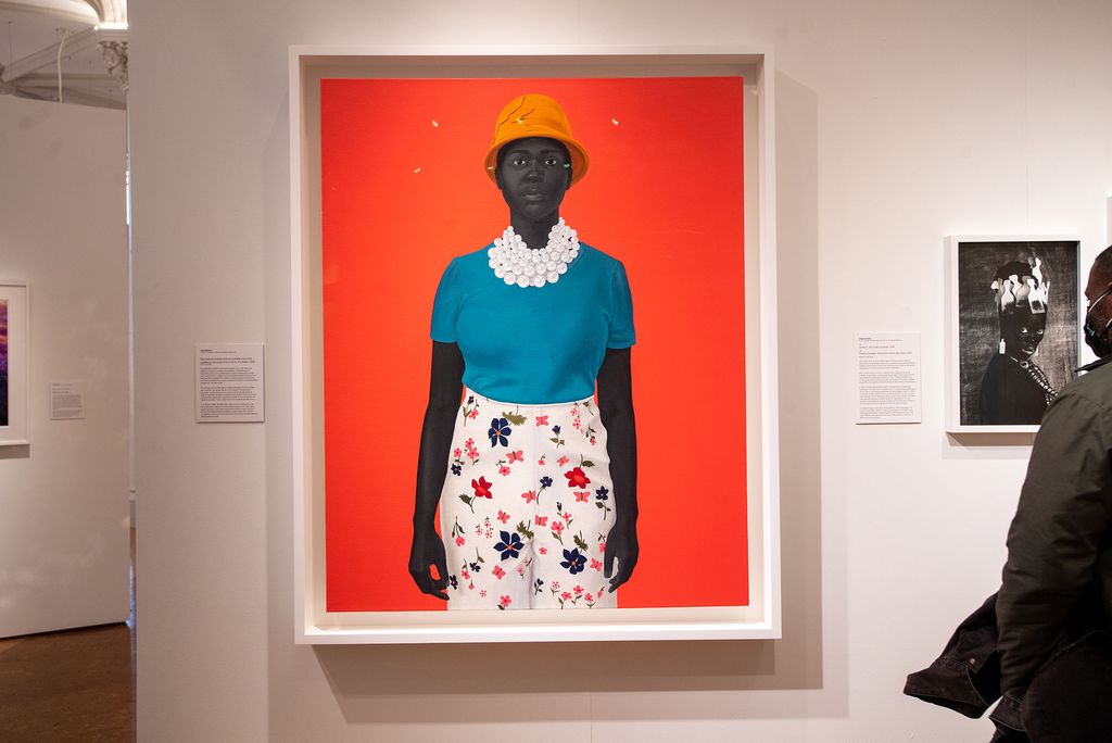 Photo of Amy Sherald’s painting "She had an inside and an outside now and suddenly she knew how not to mix them (2018)" The painting features a dark-skinned Black woman wearing an orange bowl hat, a large pearl necklace, a blue blouse, and floral white pants on a bright orange background. A gallery visitor is seen at right look at a photograph of a Black woman with white combs in her hair.