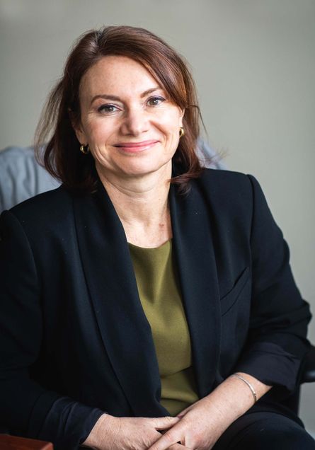 Portrait of Janna Greenberg, professor of the practice, College of Communication, sitting in a gray office chair and wearing a black blazer with a green blouse. She is a middle-aged white woman with short brown hair.