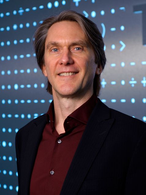 Photo of Marshall Van Alstyne, wearing a black blazer and red button-down shirt. He is a middle-aged white man with blondeish hair pushed behind his ears. He stands in front of a screen with light blue dots across it.