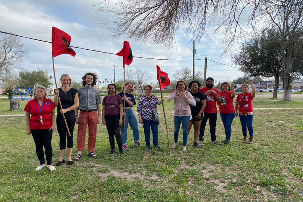 Photo: a group of Border Studies Program participants and LUPE volunteers stand in a line facing the camera near a border wall. Various people hold a red flag.