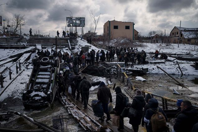 Photo of Ukrainian citizens crossing an improvised path under a destroyed bridge while fleeing Irpin on March 8. Photo shows a line of people wearing dark clothes on a dark snowy day as they follow a path near a destroyed bridge.