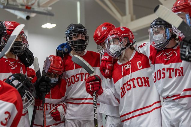Photo of the BU Roller Hockey team in a huddle. They wear red and white jerseys that read "Boston", red helmets, and hold sticks as they huddle together.
