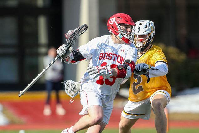 Action shot photo of Men’s lacrosse attacker Timmy Ley. A male lacrosse player wearing a protective helmet holds his lacrosse stick with the ball and defends against an opposing player in a yellow uniform.