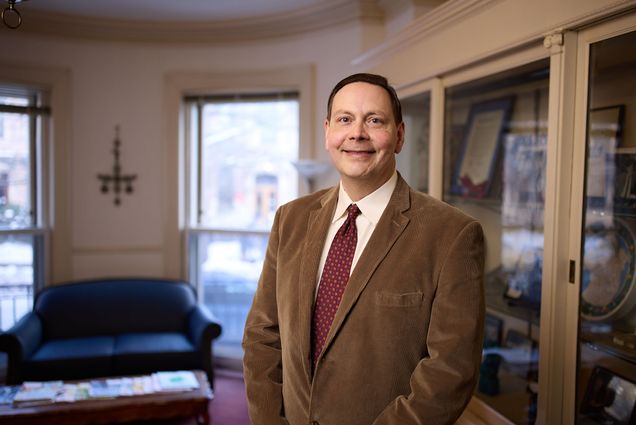 Photo of Steven Sandage, a Boston University School of Theology professor of psychology of religion and theology. He stands smiling in a white collared shirt, burgundy tie, and brown blazer. Blurred background shows a spacious office on a sunny day.