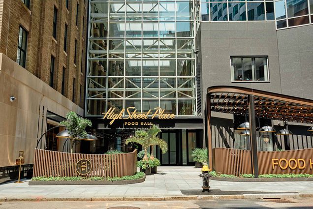 Photo of the front of High Street Food Hall. A tall, modern building surrounded by greenery and wood accents is seen with a large sign in front that reads "High Street Place, food Hall"