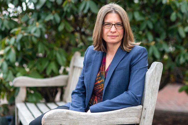 Photo of Brenda Philips sitting on a wooden bench in a park and turned to look at the camera. A white woman with glasses wearing a blue blazer, red, green, and yellow blouse, and black pants, poses for the camera.