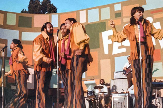 Still from the movie Summer of Soul featuring Fifth Dimension during a performance. They wear orange, yellow and brown outfits, including jackets with fringe and bell bottom pants. The women at right at the microphone raises her hands as she sings. Three men at right sing into one microphone.