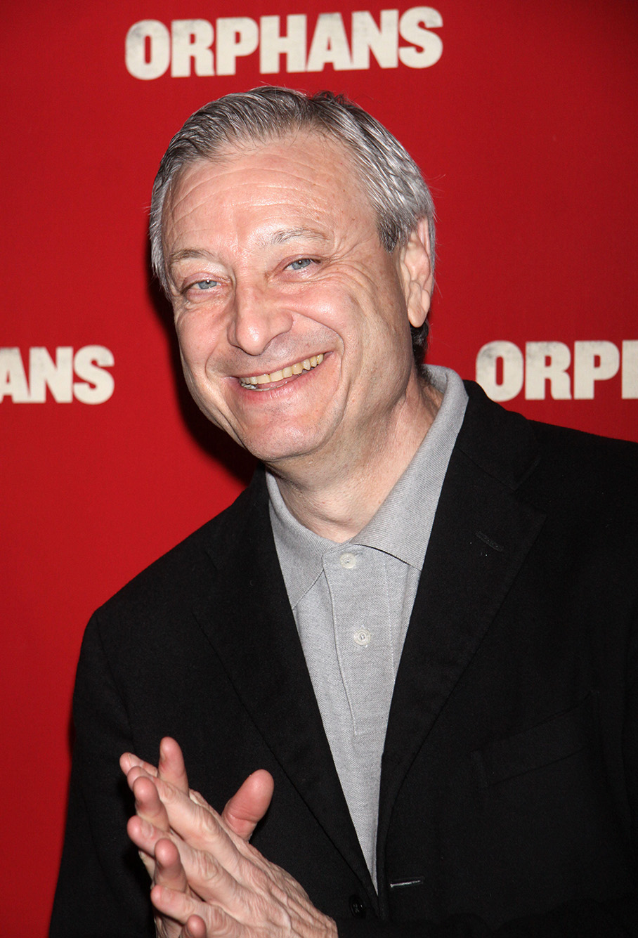 Candid photo of Fred Zollo as he stands in front of a red "Orphan" movie premiere background. An older white man with gray hair and wearing a grey shirt and black blazer smiles at the camera