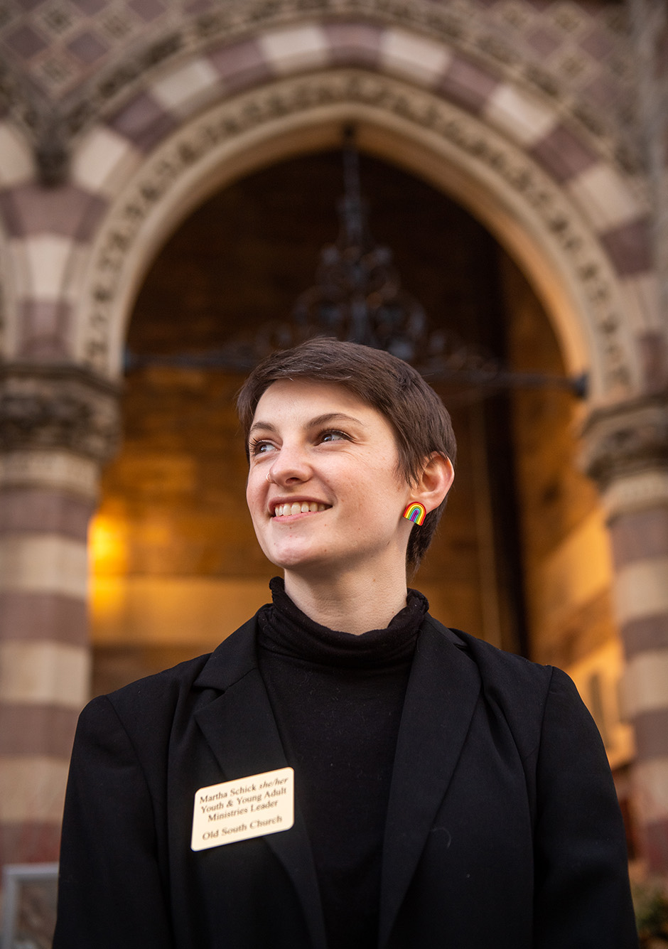 side profile photo of Martha Schick (MDiv’22). A white woman with short hair dressed in all black smiles and looks to the left. Blurred background shows the entrance to a chapel