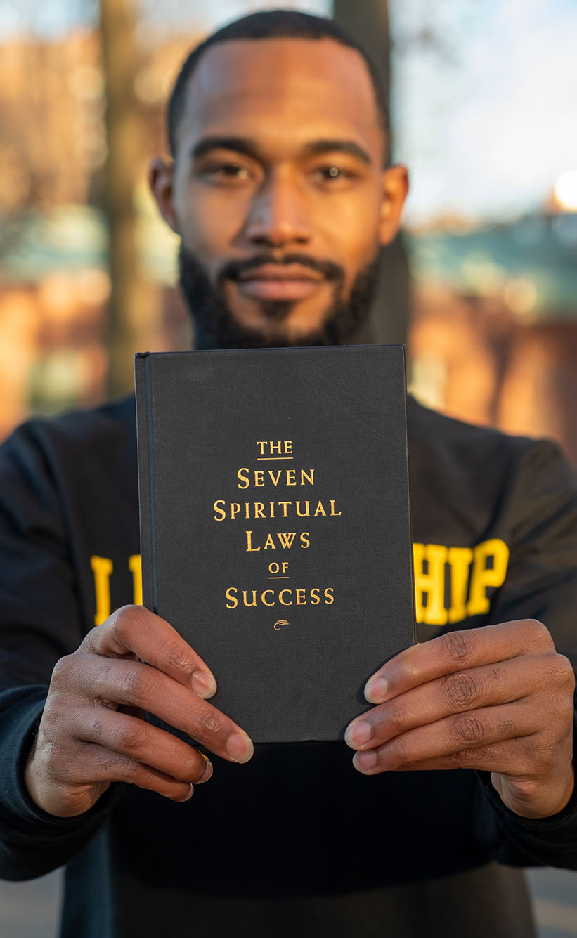 Photo of Interfaith leader Jonathan Allen, a black man with a freshly cut black hair and beard holds up in front him Deepak Chopra’s book, "The Seven Spiritual Laws of Success"