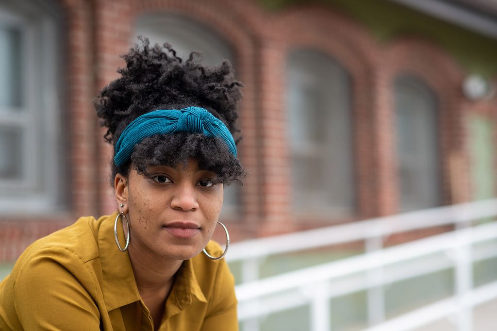 Photo of Alexis Walls (SPH’17) leaning against a while rail. She was a knotted blue headband and hoop earrings and looks earnestly at the camera. A brick building is seen in the background.