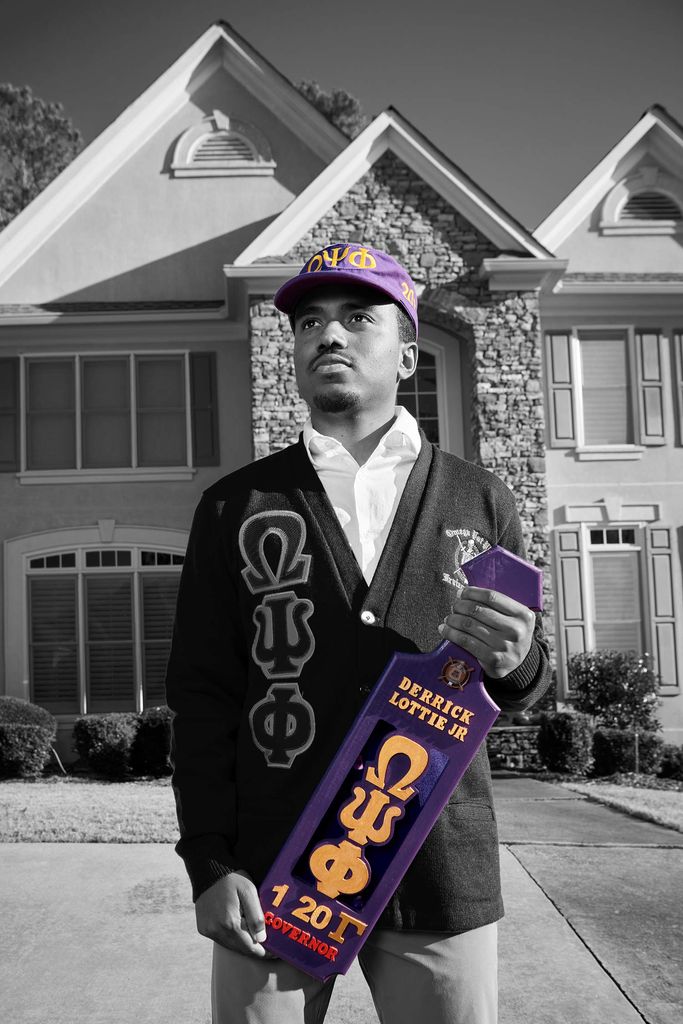 Photo of Derrick Lottie, Jr. (QUESTROM’21) standing on the lawn of a large house. The photo is black and white with the exception of his Brothers United hat and object that he holds in his hand, both are tinted a bluish purple.