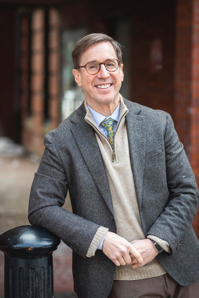 Photo of David Jernigan smiling and leaning against a post. He wears a gray suit jacket and tan quarter-zip.