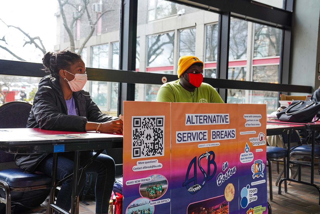 Photo of BU students Jada Peart (CAS 22'), left, and Rudy Joseph (WHEELOCK 24') sits in front of the Alternative Service Breaks table to help students to learn about alternative spring break activities during the week of spring break. Both students wear face masks and sit behind a colorful poster board with a large QR code that reads "Alternative Service Breaks"