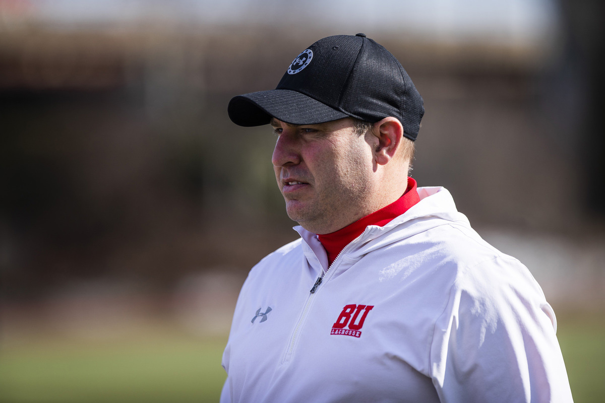 Photo of Ryan Polley, the Bu men’s lacrosse head coach. A white man wearing a black cap and white windbreaker with the Bu logo on it looks off into the distance as he watches the team.