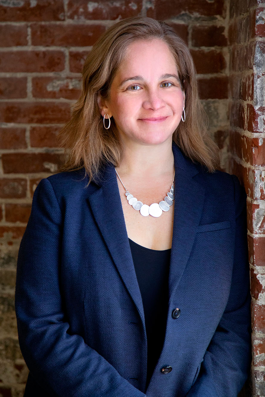 Photo of Rebecca Pearl-Martinez, a middle-aged white woman with light brown hair and wearing a black blouse and navy blue blazer. She poses against a brick wall and smiles at the camera.