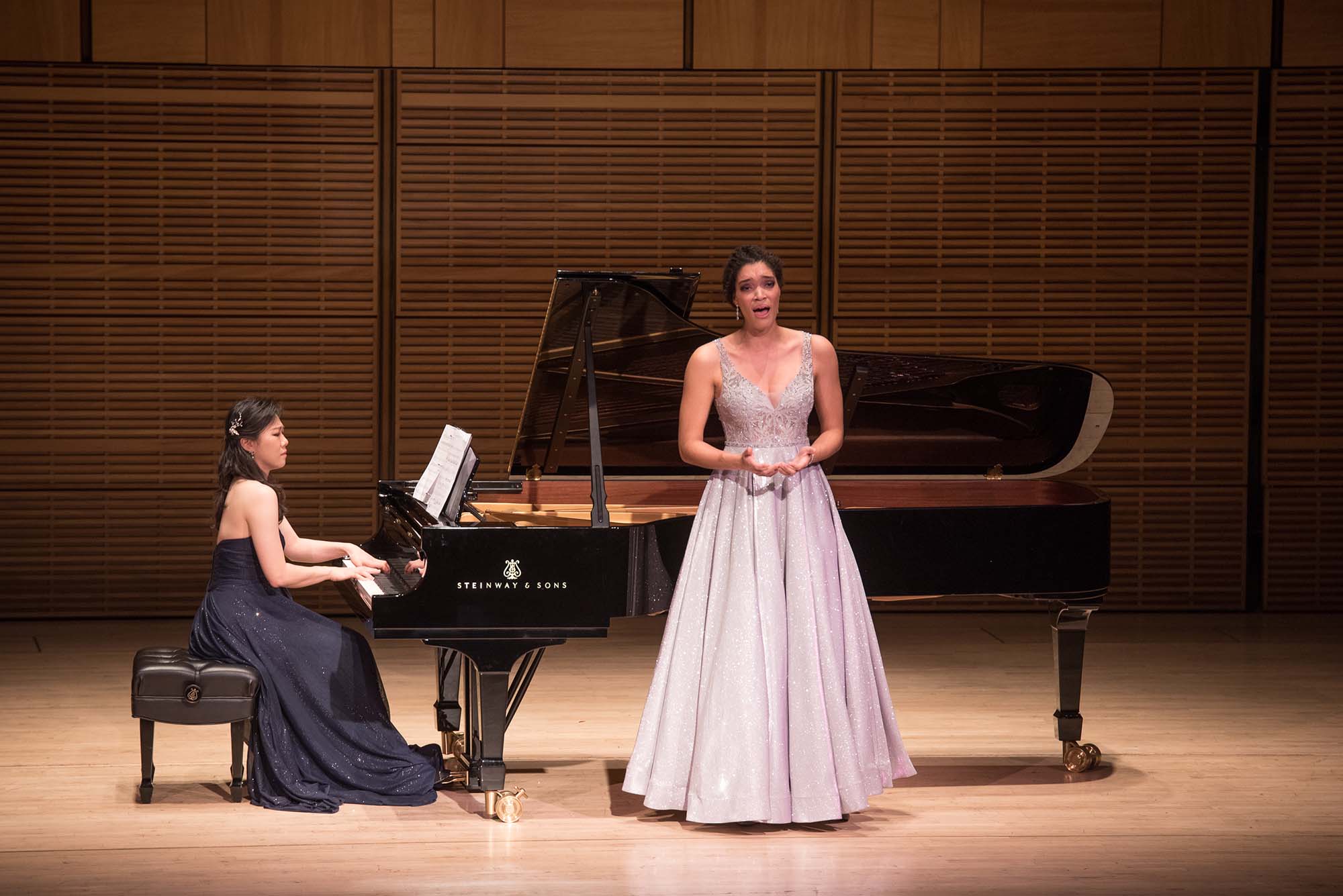 Photo of a woman of color in long, silver gown, her hands extended as she sings with a solemn look on her face. Behind her, an Asian woman in a long, sparkly, black gown plays concentratedly at a grand piano. Both women are on stage.
