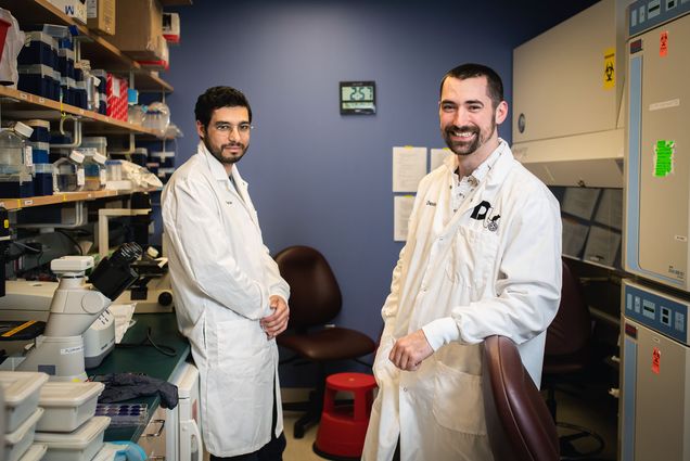 Photo of Florian Douam (left) assistant professor, microbiology, and Devin Kenney, a PhD student, posing for a photo at National Emerging Infectious Diseases Laboratories. The both wear white lab coats and stand facing each other with their bodies and heads tilted towards the camera in front of a purple-blue wall. Lab shelves can be seen on either side of them.
