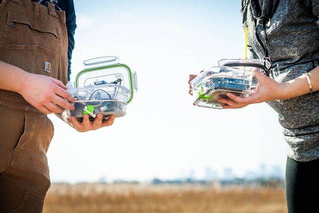 Two pairs of hands each hold a box full of scientific sensors in front of a sunlight sky background