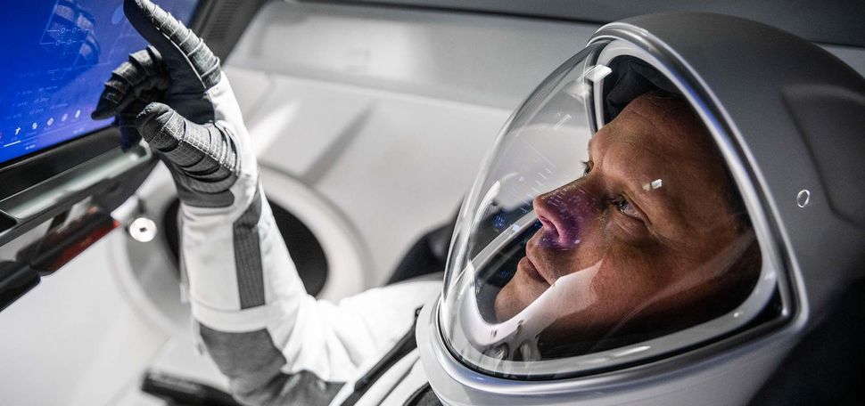 Photo of NASA pilot Bob Hines wearing a white and gray space-x suit inside during what appears to be a training drill in a space-x craft simulator. He touches a blue screen with his gloved hand and wears a sleek astronaut helmet with a glass front.