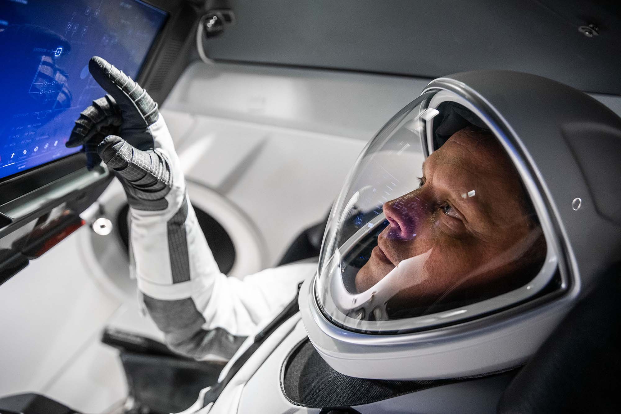 Photo of NASA pilot Bob Hines wearing a white and gray space-x suit inside during what appears to be a training drill in a space-x craft simulator. He touches a blue screen with his gloved hand and wears a sleek astronaut helmet with a glass front.