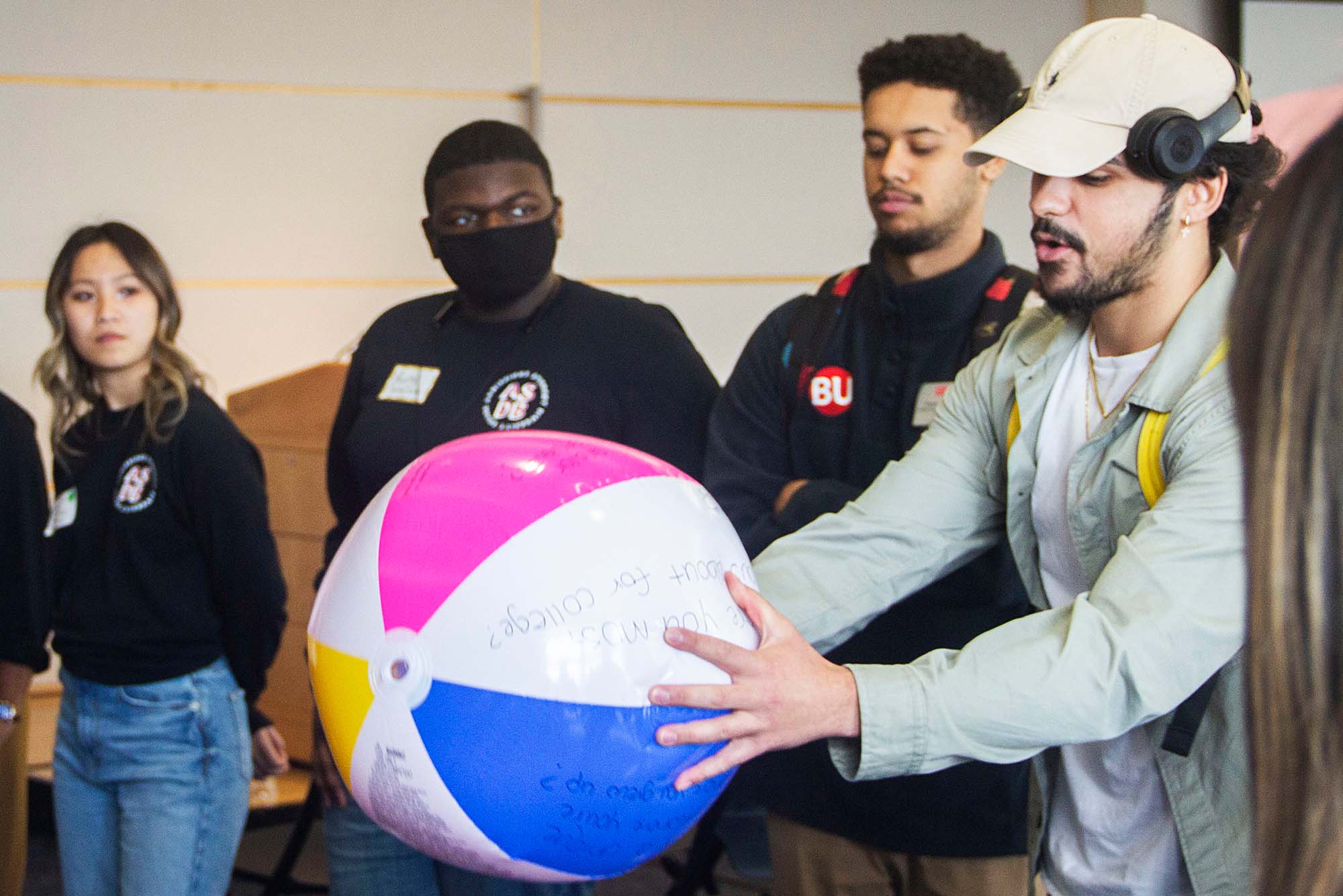 Photo of Wendell Nunes (CAS’23) catching a beach ball with questions written on it during an ice breaker activity at the Multicultural Welcome Weekend. Nunes is a brown-skinned young man who wears a white hat with headphones over it. A diverse group of students stand in a circle to his left and right.