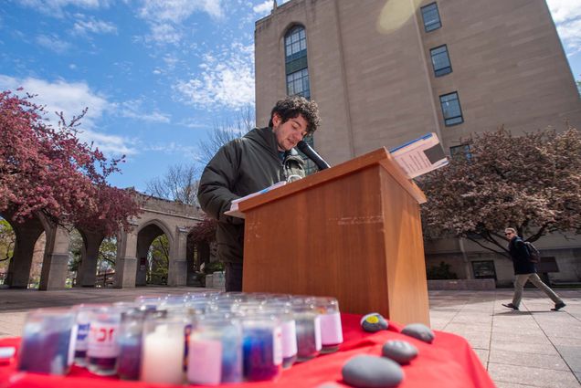 Photo of Max Rosen (Sargent’23) reading the names of victims of the Holocaust in observance of Holocaust Remembrance Day (Yom Hashoah) April 28 in Marsh Plaza. Rosen has short curly hair, wears a gray jacket, and speaks into a microphone a top a wooden podium. In the foreground, candles and small rocks are seen on a table with a red table cloth. In background, the sky is bright blue with a few clouds.