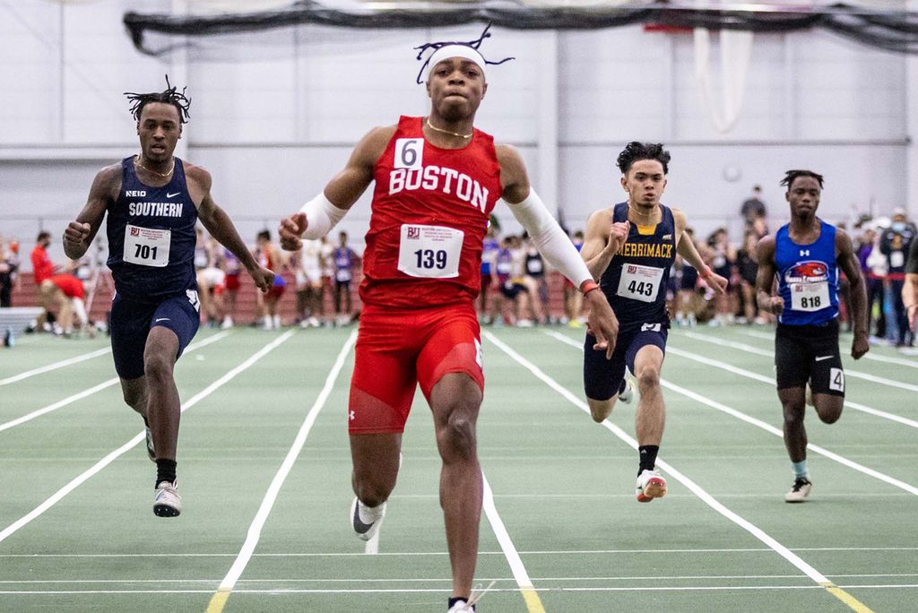 Photo of Toby Makoyawo, a young Black man wearing a red "Boston" track jersey and long red spandex shorts, is shown mid-stride next to a young Black man in a dark blue track uniform that reads "Southern", a young man with a dark blue and yellow track uniform that reads "Merrimack", and a young man with a blue and red track uniform; all are also mid-stride. They all are running towards the camera down the straightaway of the track of the green indoor track of the BU Track and Tennis Center. The starting blocks and various other athletes and coaches can be seen in the blurry background.