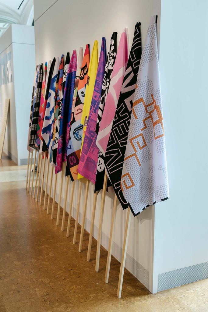 Photo of a series of colorful flags with geometric patterns lined up against a white wall.
