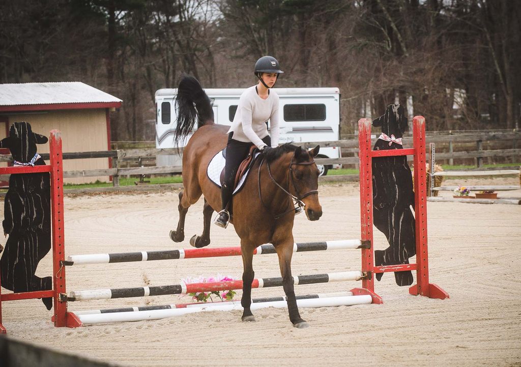 Action photo of BU Equestrian Team cocaptain Lizzy Nevins in white riding gear and black helmet riding a brown horse with a black mane and jumping over barriers in a sandy horse ring.