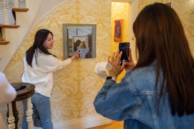 Photo of Heeseung “Chelsea” Han (CFA’23), left, posing with her piece “Back to Me” while her friend Mina Chung (CFA’23) snaps a photo, right foreground, during the during the biannual art exhibit at Sloane House April 11. Han holds a pen and pretends to draw on her piece, which is an image of a person painting a boston skyline painting a boston skyline. The house has ornate, gold and white floral wallpaper. Han is a young, Asian woman with long dark hair and wears a white sweater and jeans.