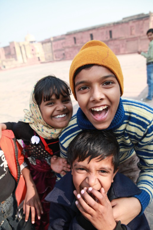 Photo of three Indian children smiling and laughing excitedly for the camera. One small child covers their mouth, one wears a yellow cap and striped green and blue shirt, and one wears a pastel yellow scarf, black long-sleeved shirt, and pink dress.