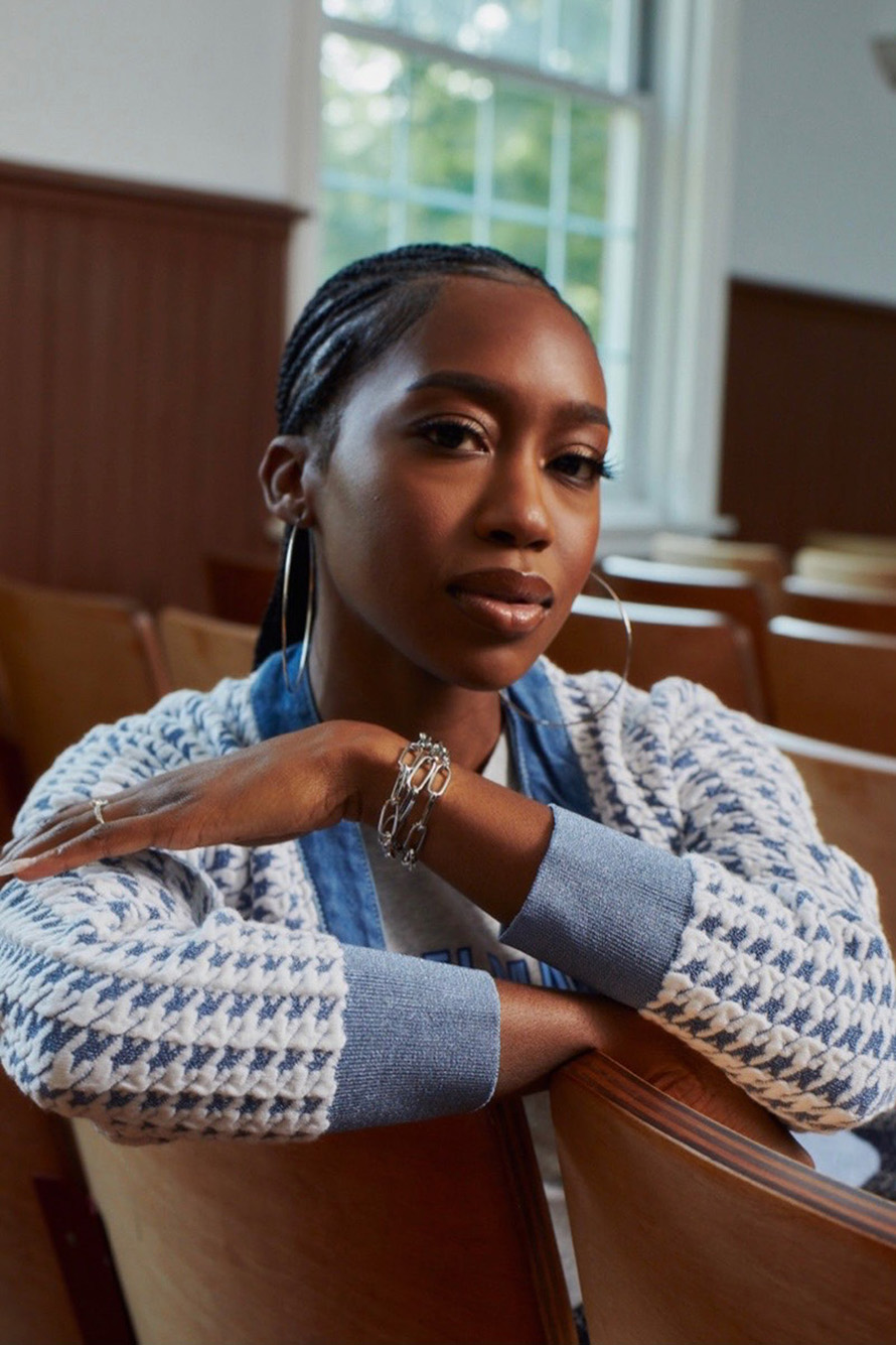 Photo of Lynae Vanee Bogues sitting in what looks to be a church or auditorium. A young black woman with neat cornrows and a light blue cardigan is turned in her seat with arms resting on the head of her chair as she poses for the camera.