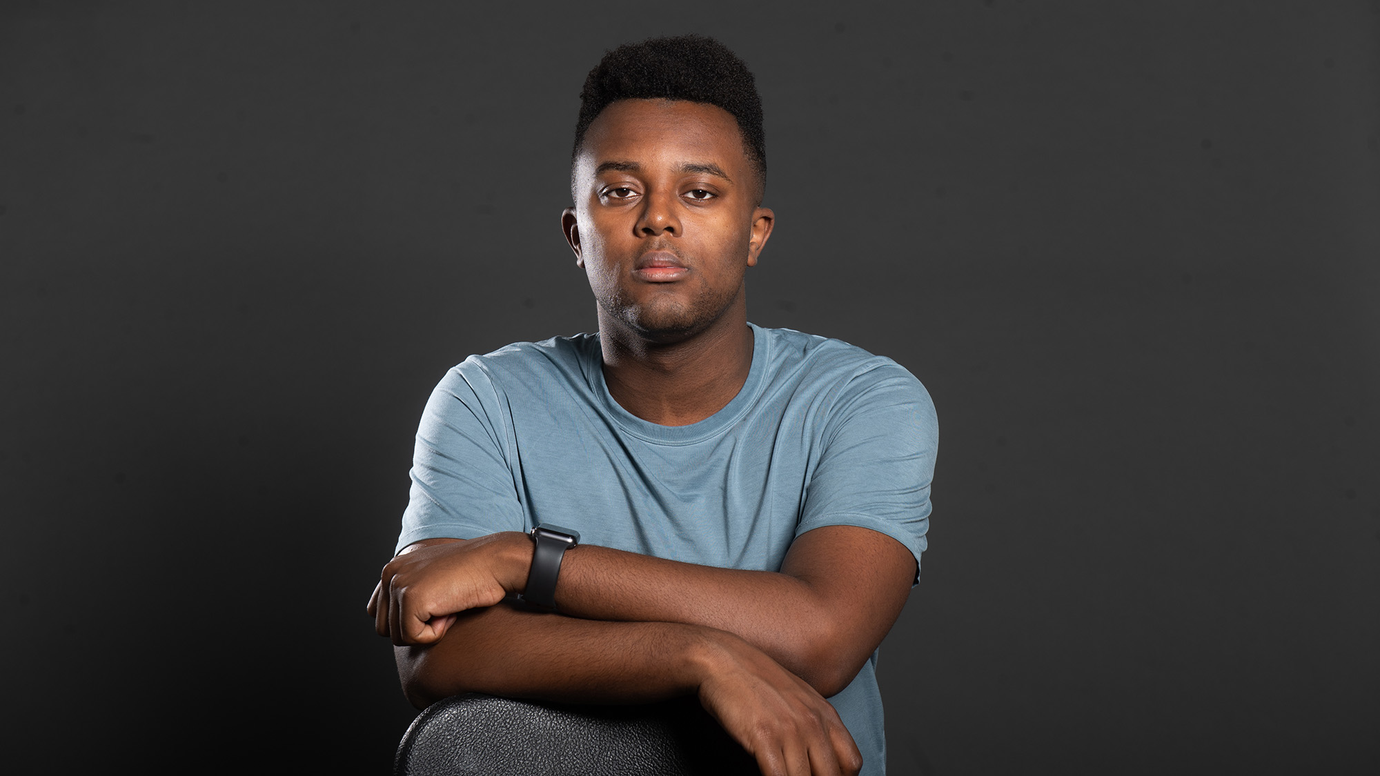 Studio portrait of Amos Mwaura. A young black man with a short afro and somber expression leans on a chair with arms crossed on top of a black chair. He wears a light blue, short-sleeved t-shirt.