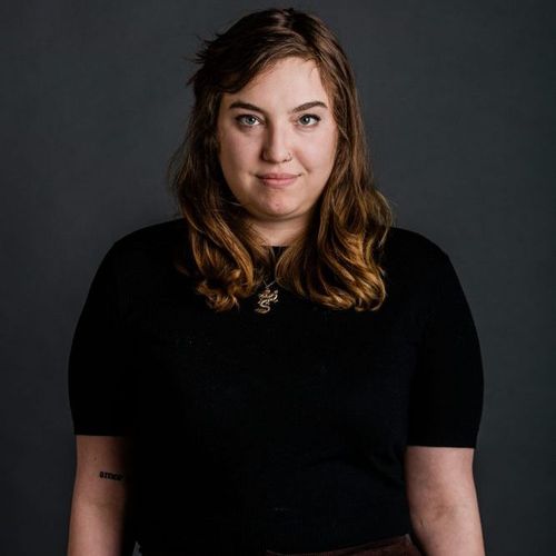Photo: Headshot of Sophie Yarin. A white woman with wavy brown hair and wearing a black dress and gold necklace, smiles and poses in front of a dark grey backdrop.
