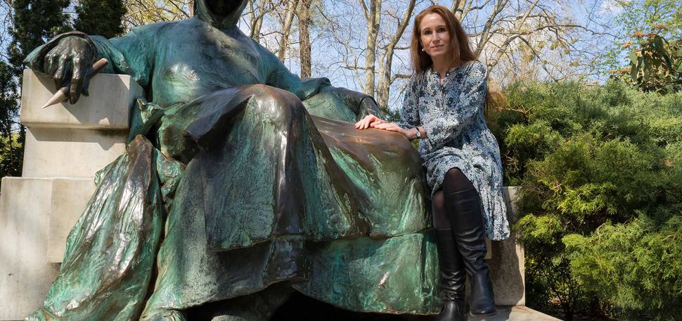 Photo of Theodora Goss in Budapest park with teal rusting bronze statue of Anonymus, a well known landmark. A white and red hot air balloon can be seen in the distance of the background, behind naked tree branches.