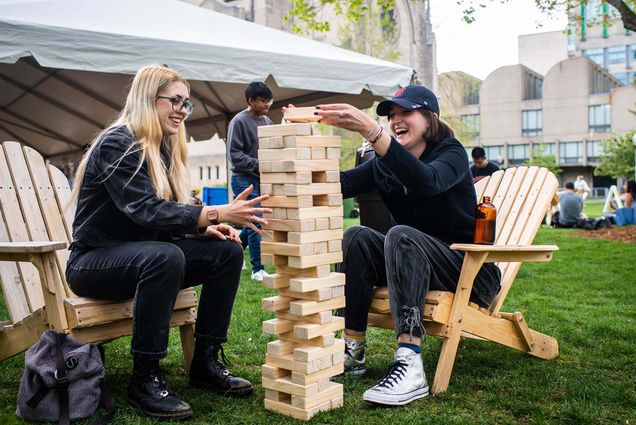 Photo of Eden McColl (SHA 21) and Morgan Michelson (SHA 21) playing a game of Jenga during Senior Week. The pair sit on wooden adirondack chairs, as McColl holds the tower from tipping over and Michelson attempts to place a piece on top. Both students wear black, Michelson wears a Red Sox hat. Behind them, a white tent and other students are seen on the BU beach.