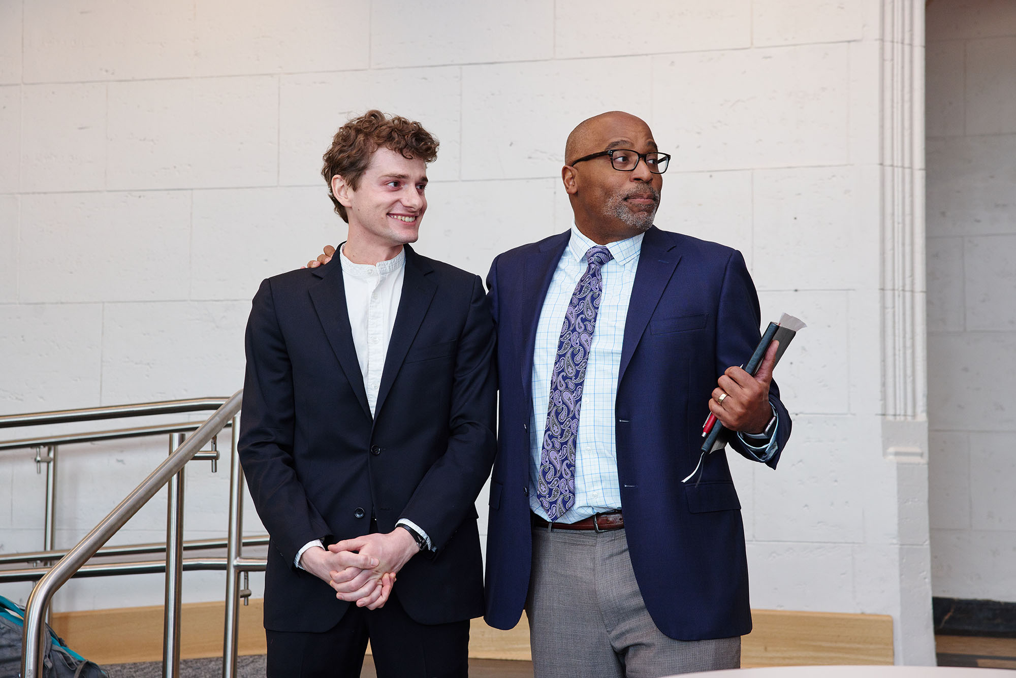 Photo of Painter and wood sculptor Gavin Fahey (CFA’22) standing to the left of Harvey Young, College of Fine Arts dean. A young white man wearing a white shirt, black blazer, and black pants stands next to an older Black man wearing a navy blazer and grey pants. They both look to the left in this candid shot.