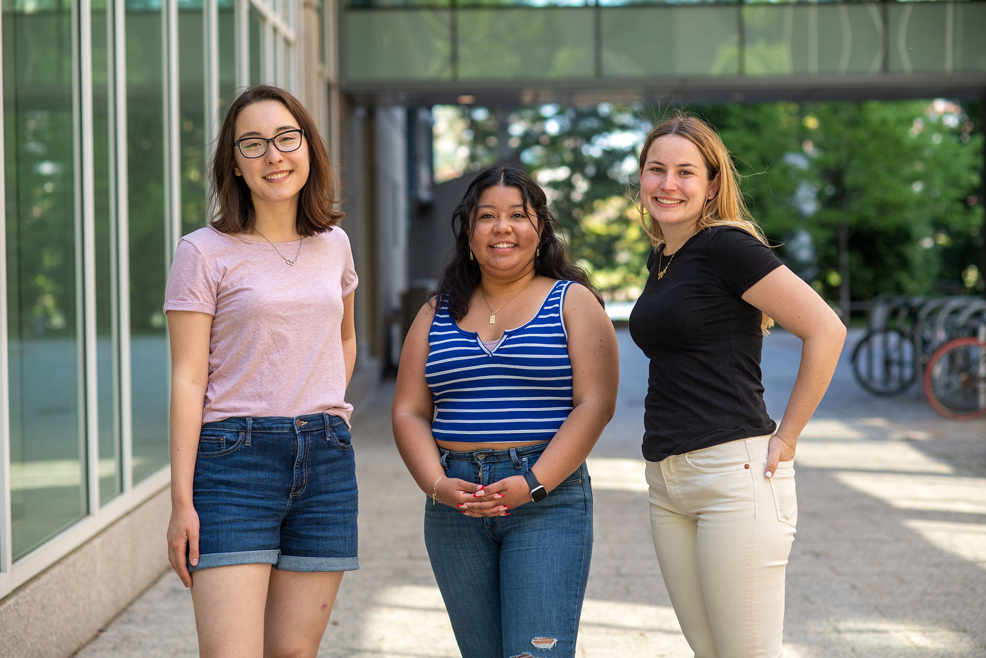 Photo of Newbury center grads Maelee Chen, from left, Cecilia Jarquin, and Annie Rose Hallett. Chen wears a pink tee shirt and jean short, Jarquin wears a striped blue and white tank top and blue jeans, and Hallett wears a black shirt and light tan pants. All 3 young women stand together and pose for the camera.