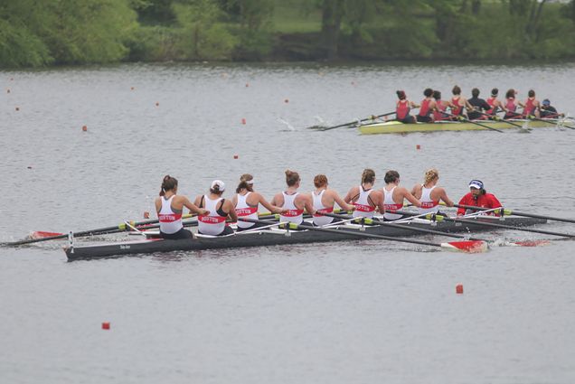 Photo of two rowing boats on a river during an overcast day. One boat is full of rowers from the back wearing white tops, the other further in the background shows rowers wearing red.