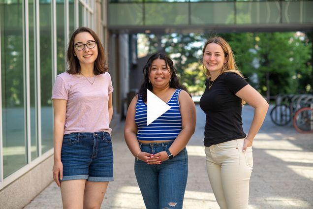 Photo of Newbury center grads Maelee Chen, from left, Cecilia Jarquin, and Annie Rose Hallett. Chen wears a pink tee shirt and jean short, Jarquin wears a striped blue and white tank top and blue jeans, and Hallett wears a black shirt and light tan pants. All 3 young women stand together and pose for the camera. A white triangle play button is overlaid in the middle of the photo.