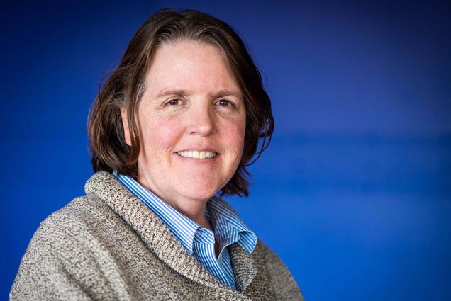 Photo of Metcalf winner Leslie Dietiker, an older white woman with short brown hair with some gray streaks in it. She smiles, and wears a thick, gray sweater and striped blue shirt. She stands in front of a blue background.
